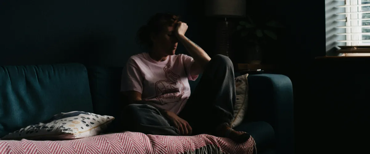 Person sitting alone in a dark room with head in hand, symbolising the loneliness, exhaustion and emotional struggle of living with addiction