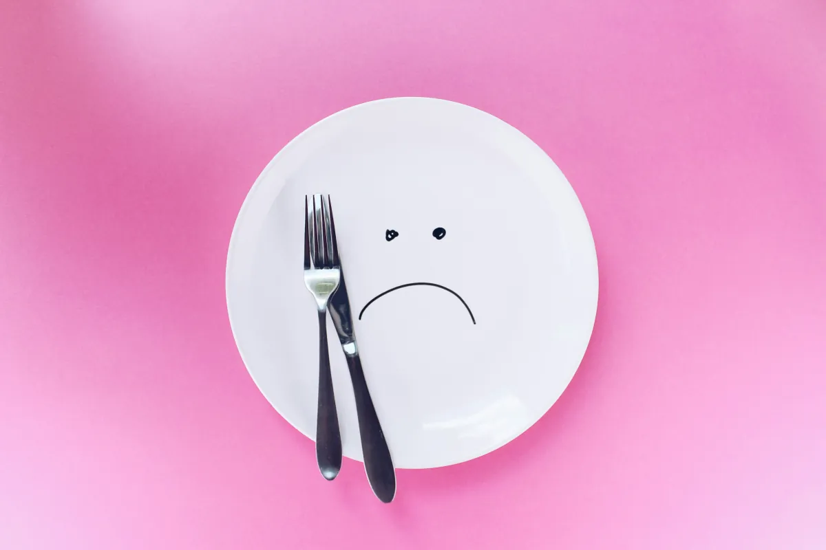 Empty white plate with a sad face and cutlery on a pink background, symbolising the emotional pain and struggle of living with an eating disorder