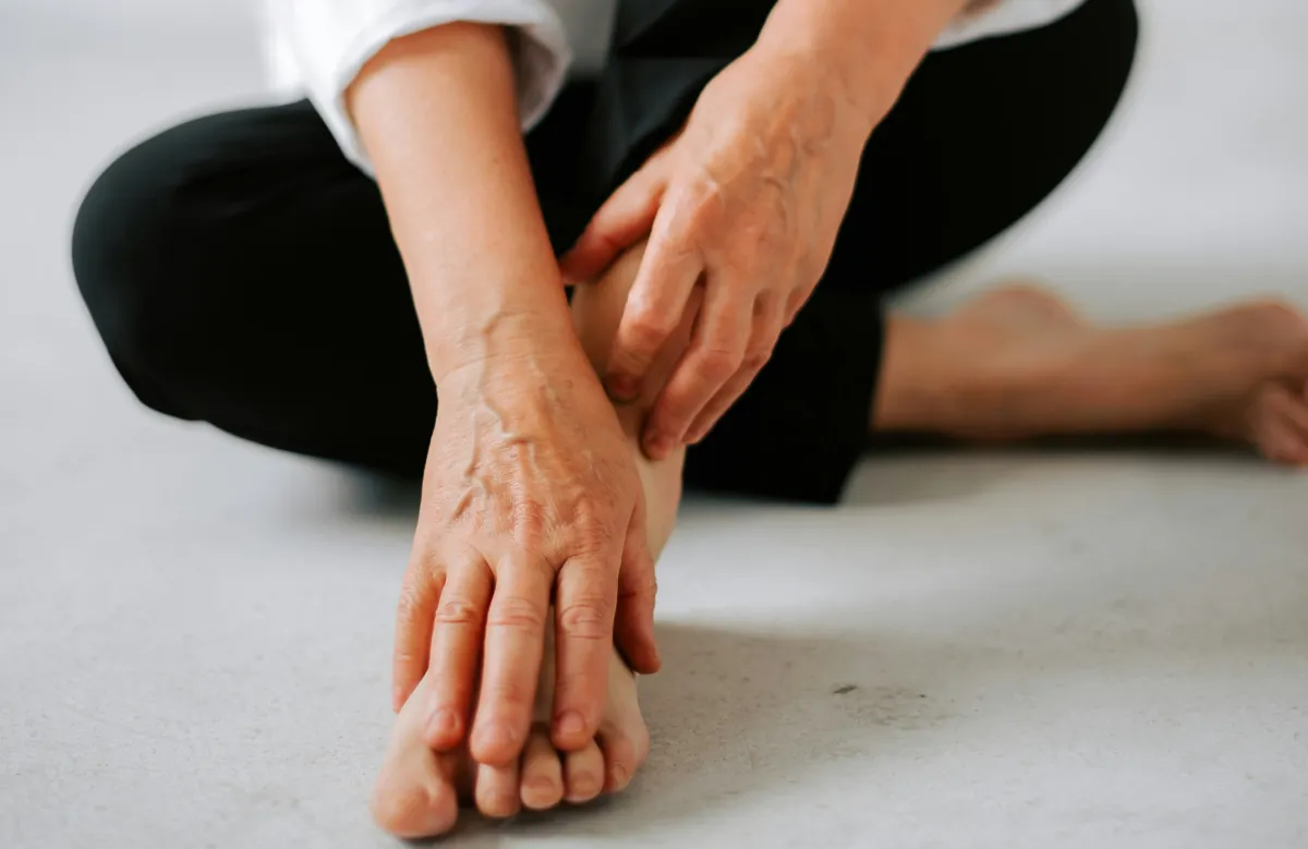 Middle-aged woman sitting barefoot on the floor gently holding her foot, symbolising the physical discomfort and emotional challenges of perimenopause and menopause