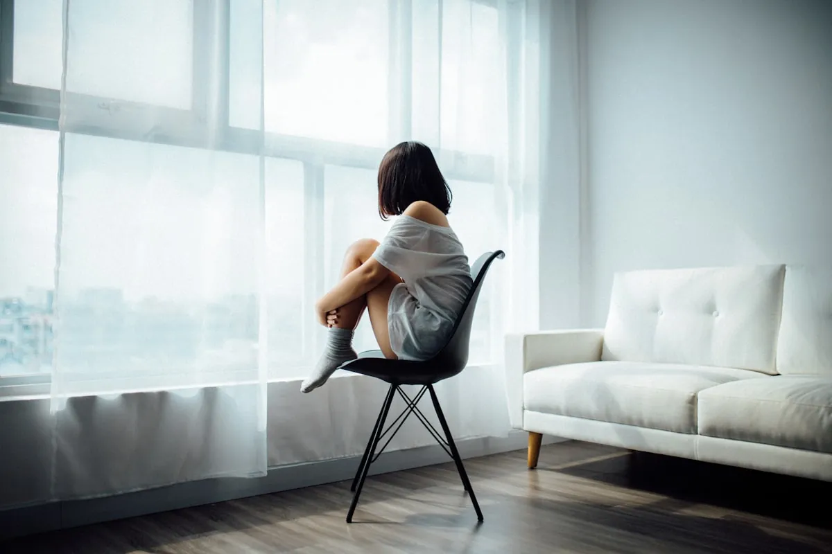 Young woman sitting alone on a chair gazing out of a window, symbolising loneliness, self-doubt and the silent struggle of low self-worth and inner criticism