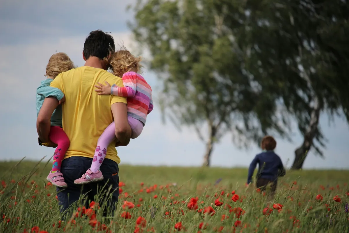 Single father carrying two young children through a blooming poppy field while a third child runs ahead, symbolising the love, strength and daily challenges of single fatherhood